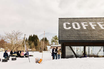 Une scène extérieure enneigée présente un café moderne avec "COFFEE" sur le toit. Les gens se rassemblent à l'extérieur, socialisant et sirotant des boissons, entourés d'arbres et de montagnes.