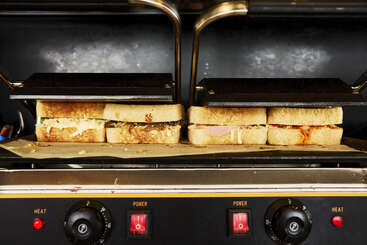 Quatre sandwichs sont grillés dans une presse à panini commerciale, le pain doré et les garnitures visibles, sous des plaques métalliques chaudes, prêts à être servis dans une cuisine.