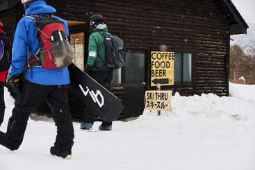 Deux snowboarders marchent dans la neige en direction d'un bâtiment rustique en bois. Des panneaux annoncent du café, de la nourriture, de la bière et la possibilité de faire du ski, avec une écriture japonaise en dessous. Scène hivernale.