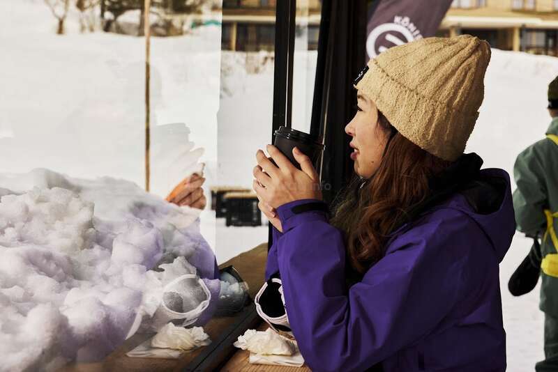 A woman in a purple jacket and beige beanie enjoys a hot drink by a snowy window, her reflection visible, creating a cozy winter atmosphere indoors.