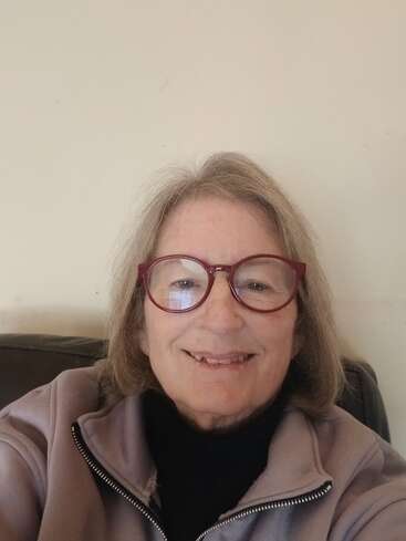 Une femme âgée souriante, aux cheveux gris mi-longs et aux grosses lunettes à monture rouge, est assise à l'intérieur, vêtue d'une veste légère et d'un haut noir, sur un fond uni.