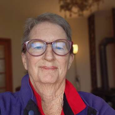 An older woman with short gray hair and large glasses smiles gently. She wears a red-collared shirt and purple jacket. The background is softly lit indoors.