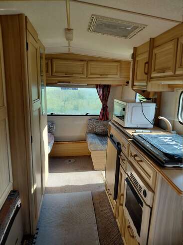 This image shows the interior of a cozy camper van featuring a compact kitchen, wooden cabinets, oven, a microwave, seating area, window, and storage compartments.