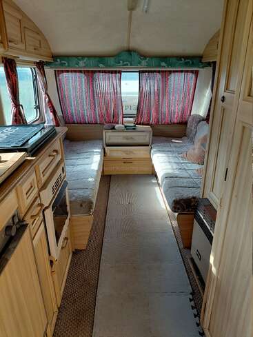 This image shows the cozy interior of a camper van or caravan with two cushioned benches, a small kitchen, wooden cabinets, and colorful patterned curtains.