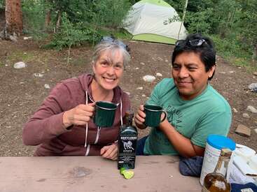 Das Bild zeigt ein Paar, das an einem Picknicktisch in einem Waldgebiet sitzt, jeder mit einem Becher in der Hand, umgeben von Campingausrüstung und einer Flasche Montelobos Espadin Mezcal.