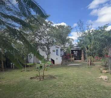 A imagem mostra uma casa branca serena com um quintal exuberante, cercada por árvores e um céu azul com nuvens, exalando uma atmosfera pacífica e natural.
