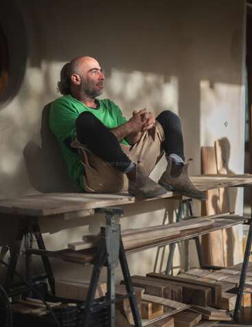 Ein Typ in einem grünen Hemd und Stiefeln sitzt nachdenklich auf einer Werkbank, umgeben von Holzplanken, während Sonnenlicht Schatten an die Wand hinter ihm wirft.