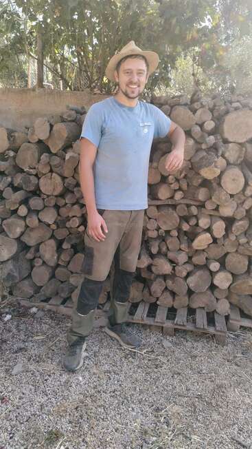 A smiling man in a hat stands in front of a large stack of firewood outdoors. He wears work clothes and boots, surrounded by trees and sunlight.