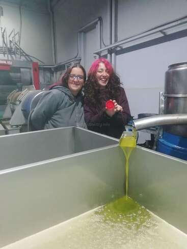 Two smiling women stand in an industrial setting, watching fresh olive oil pour from a spout into a large metal container. One holds a red cap.