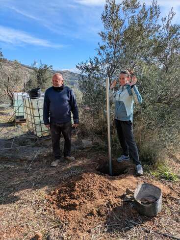 Deux personnes se tiennent à l'extérieur, près d'un trou fraîchement creusé et entouré d'arbres. L'une sourit, l'autre lève les mains, tenant un poteau. Paysage rural ensoleillé, ciel bleu.