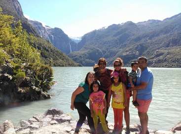 La imagen muestra a una familia de ocho miembros posando para una foto sobre unas rocas junto a un lago, con montañas al fondo y una cascada visible a lo lejos.