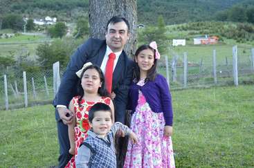 La imagen muestra a un hombre trajeado posando con tres niños en un campo cubierto de hierba, con un árbol y una valla al fondo.