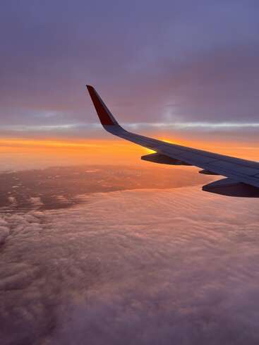 La imagen capta el ala de un avión elevándose por encima de nubes esponjosas con una impresionante puesta de sol naranja y morada, creando una vista tranquila y de ensueño desde el asiento de la ventanilla.