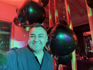 Un hombre sonriente se toma un selfie en una fiesta con globos negros en forma de corazón de fondo, iluminada por luces de colores y decoraciones festivas en la sala.