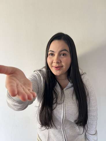Una joven de pelo largo y oscuro, vestida con una sudadera gris claro, tiende la mano hacia la cámara, sonriendo cálidamente, de pie contra una pared lisa.