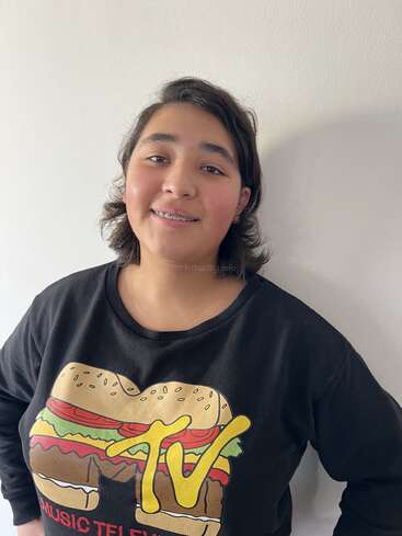 Un joven sonriente con el pelo corto y aparatos ortopédicos está de pie contra una pared lisa, llevando una camiseta negra con un gráfico de hamburguesa y el logotipo de MTV. Ambiente informal y alegre.