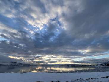 Un cielo espectacular lleno de nubes gruesas de color gris azulado se refleja en un lago tranquilo y helado. La nieve cubre el suelo, creando un paisaje invernal tranquilo e hipnotizador.