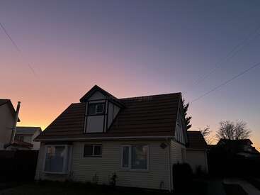 Una acogedora casa con tejado a dos aguas se recorta contra un hermoso y colorido cielo al atardecer. A su alrededor se ven casas vecinas y árboles desnudos. Atardecer apacible.