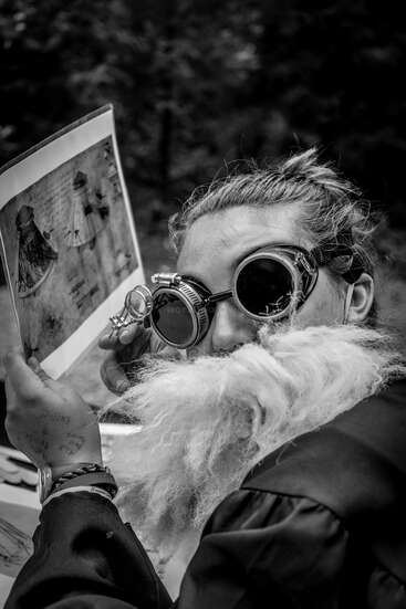 A imagem mostra uma pessoa com uma longa barba branca, usando óculos de proteção e segurando um livro, em um fundo externo desfocado.