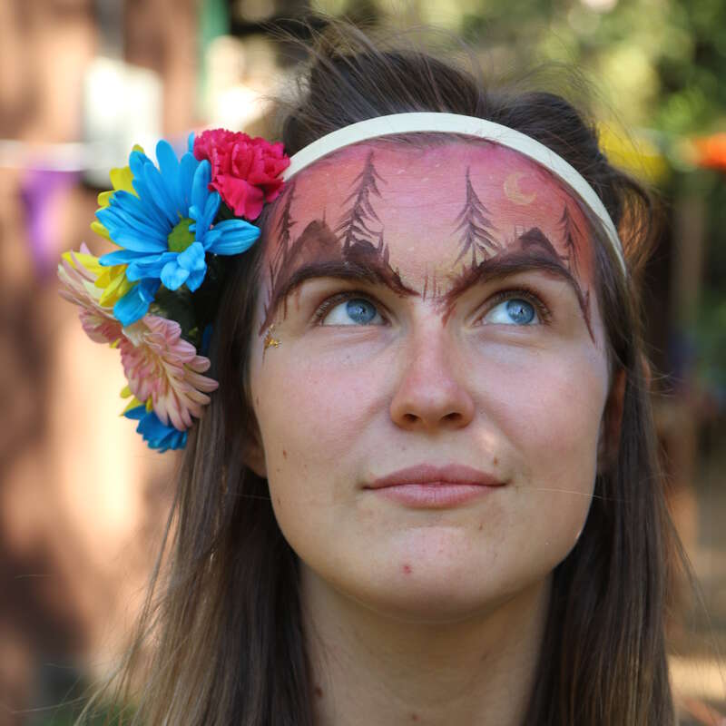 A imagem mostra uma mulher com longos cabelos castanhos, adornada com uma faixa branca e flores vibrantes, com uma pintura facial de árvores e uma lua crescente na testa.