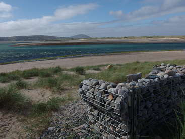 The image depicts a serene beach scene with a metal cage filled with rocks in the foreground, set against a backdrop of a sandy shore and a body of water.