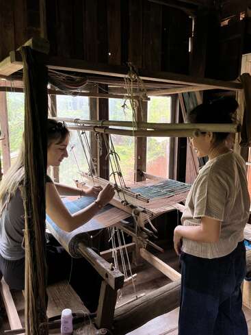 Deux femmes se trouvent dans une pièce en bois. L'une tisse du tissu sur un métier à tisser traditionnel, tandis que l'autre observe, peut-être en donnant des instructions ou en guidant le processus de tissage.