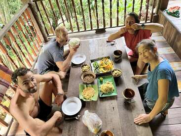 Quatre amis sont assis autour d'une table rustique en bois et dégustent un repas coloré composé de riz, de légumes et de boissons, au milieu d'une végétation luxuriante, sous un porche en plein air.