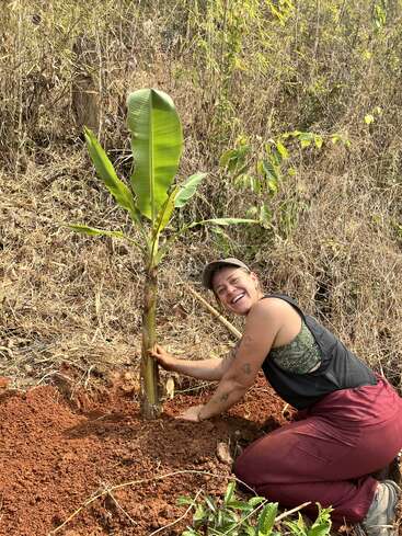 Une personne souriante est agenouillée sur un sol rouge, plantant un jeune bananier. Autour d'elle, de l'herbe sèche et du feuillage, ce qui témoigne d'un engagement en faveur de la conservation et de la croissance de l'environnement.