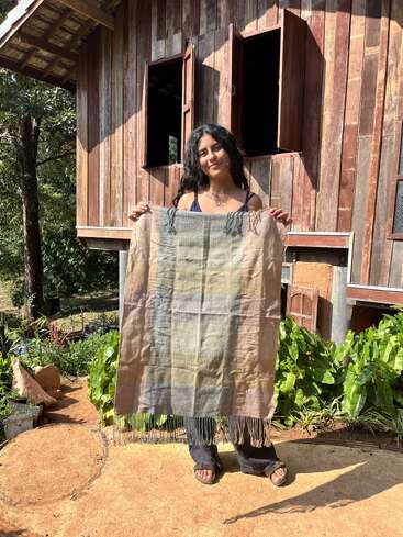 Une femme se tient à l'extérieur d'une maison rustique en bois, souriante et tenant un textile tissé à la main avec des bords frangés. La lumière du soleil et la verdure l'entourent, créant une scène paisible.