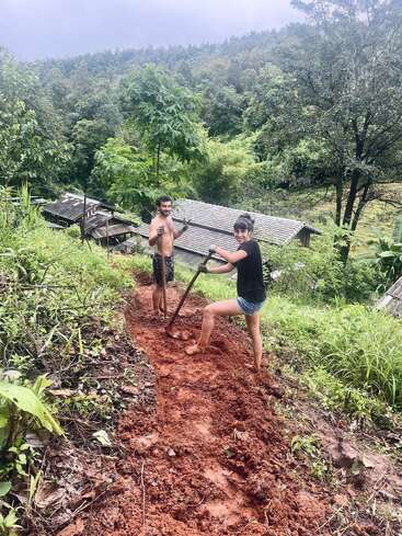 Duas pessoas estão cavando um caminho lamacento em uma colina verdejante. Elas estão sorrindo, cercadas por árvores e casas rústicas de madeira, com o céu nublado acima.