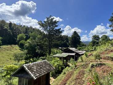 Esta imagem mostra uma paisagem rural tranquila com casas de madeira, árvores verdes, campos em terraços e um céu azul brilhante cheio de nuvens brancas e fofas.