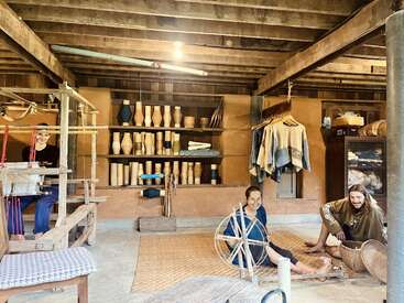 Tres personas trabajan en un rústico taller de tejido tradicional con vigas de madera. En las estanterías se exponen cerámicas, carretes y tejidos. El ambiente es cálido, artesanal y acogedor.