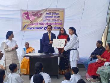 A girl in a karate uniform receives a certificate from a man, while others look on. The event is celebrating International Women's Day in India.