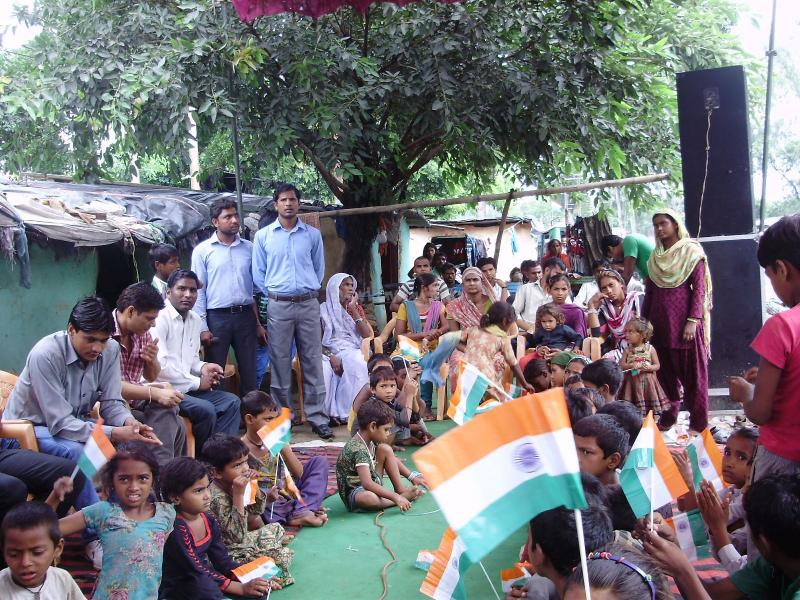 The image depicts a group of people, including children, gathered in an outdoor setting, surrounded by trees and a makeshift structure, holding Indian flags.
