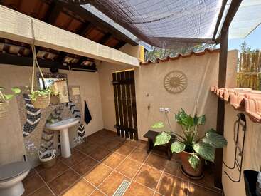 This image shows a rustic, open-air bathroom with tiled floors, a sink, toilet, potted and hanging plants, mosaic wall, wooden accents, and bamboo shading overhead.