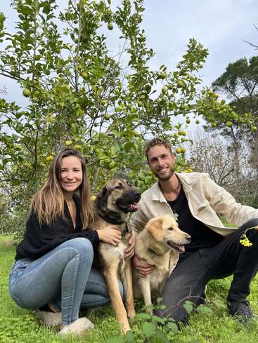 L'image représente un jeune couple posant avec leurs chiens dans une zone herbeuse luxuriante, entourée d'arbres et de buissons.