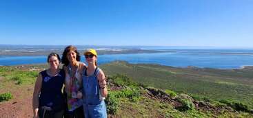 Tres mujeres están juntas sonriendo en una colina cubierta de hierba, contemplando un pintoresco paisaje costero con agua azul, tierra verde y un cielo despejado y soleado.