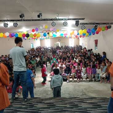 The image depicts a large room filled with children and adults, featuring a stage, colorful balloons, and a crowd of people gathered on the floor and seated on benches.