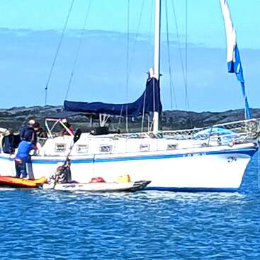 Un velero blanco está anclado en el agua con dos personas cerca. Hay kayaks junto a él, y al fondo se ve la costa rocosa y el cielo azul.