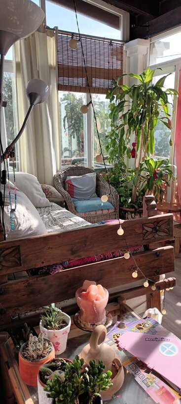 A cozy sunroom filled with natural light, plants, wicker chair, wooden sofa, string lights, small potted succulents, salt lamp, and crafts create a peaceful atmosphere.