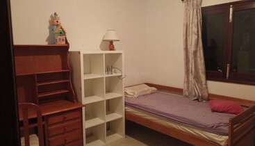 This cozy bedroom features a single wooden bed with purple bedding, a white shelf, wooden desk, chair, lamp, toy house, and a window with curtains.