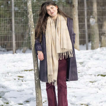 A young woman with long hair leans against a tree in a snowy park. She wears a long coat, burgundy pants, and a beige scarf, smiling gently.