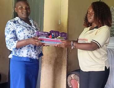 Two women are standing indoors; one is handing a bundle of notebooks, pens, and sanitary pads to the other, symbolizing support, donation, and community assistance.
