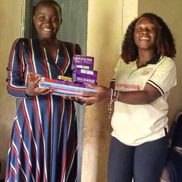 Two women are smiling indoors. One is handing over a pile of books, pens, and sanitary pads to the other, showing a moment of support and kindness.