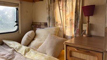 This cozy camper interior features a neatly made bed with multiple pillows, patterned curtains, a window, wooden nightstand, and a red lampshade, welcoming natural light inside.