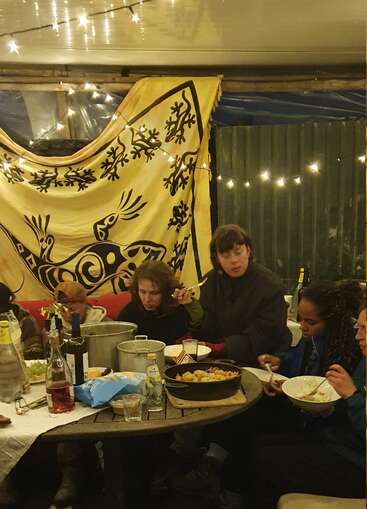 A group of people share a cozy meal, seated around a table with food and drinks, under string lights and a decorative yellow tapestry featuring a lizard.