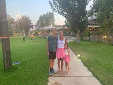 A woman and two children stand on a path in a park, surrounded by green grass and trees. Restaurant and outdoor seating are visible behind them.