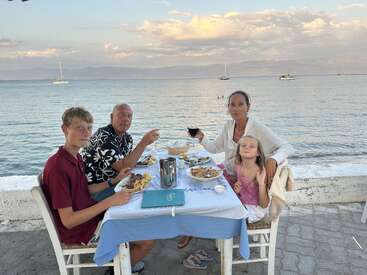 A family of four enjoys a seaside dinner at a table with pasta and drinks, with calm water, boats, and mountains beautifully in the background.