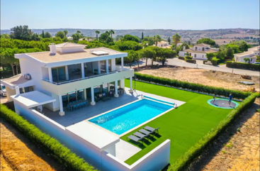 Modern two-story villa with a large swimming pool, sun loungers, spacious patio, manicured lawn, trampoline, surrounding greenery, and scenic distant views under bright sunlight.