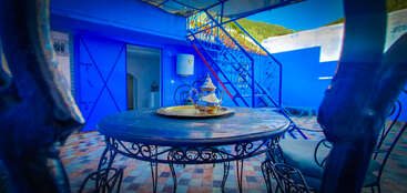 Un patio confortable avec des murs bleus et des meubles en métal. Une table circulaire accueille une théière traditionnelle. La lumière du soleil éclaire l'atmosphère vibrante et sereine. Escalier visible.